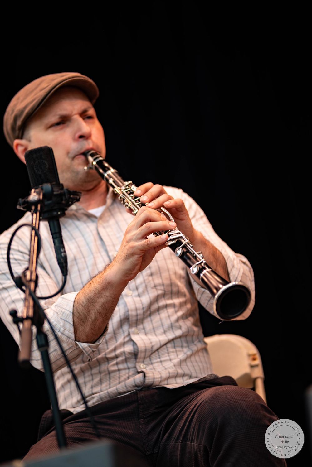 Man in a plaid shirt and cap playing clarinet on stage near a microphone.