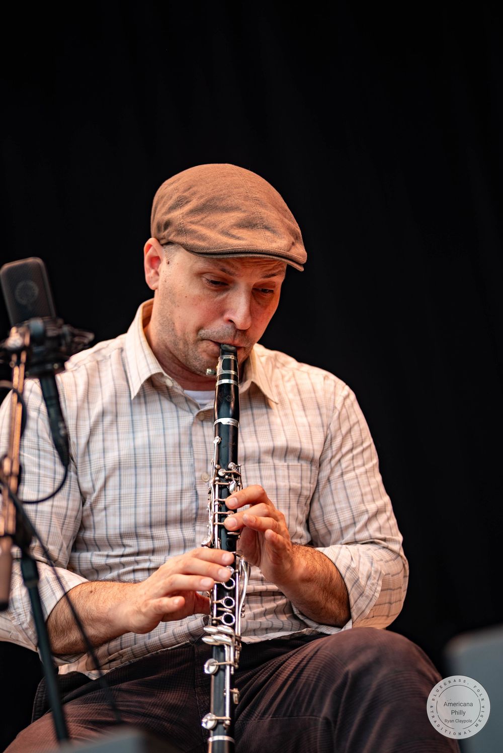 A focused male musician plays clarinet on stage, wearing a brown flat cap and plaid shirt.