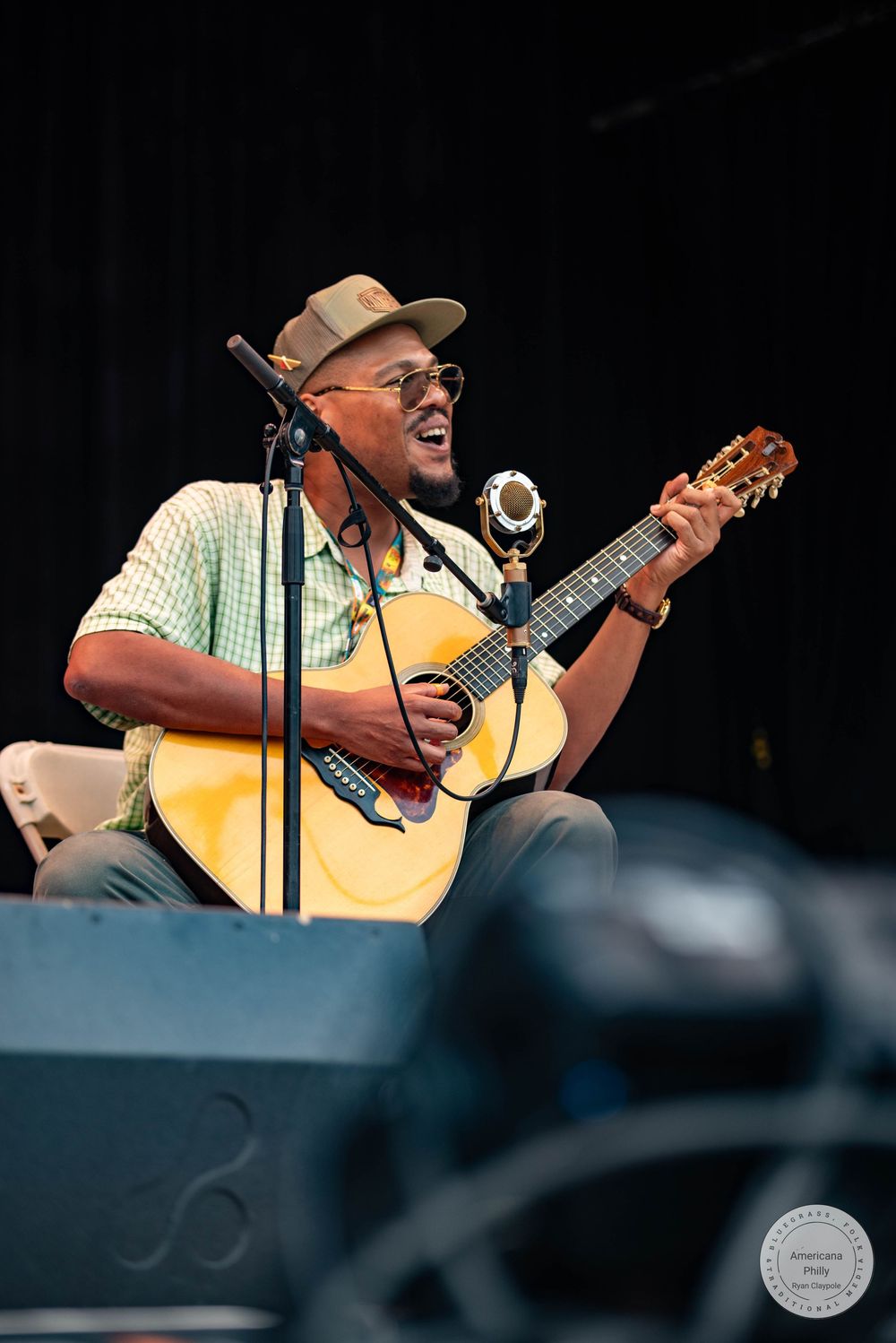 Smiling Black musician plays acoustic guitar on stage, wearing a hat and sunglasses, performing at an outdoor music festival.