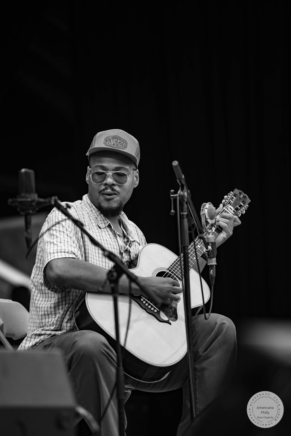 Black and white photo of a smiling musician playing acoustic guitar on stage, wearing a cap and glasses.