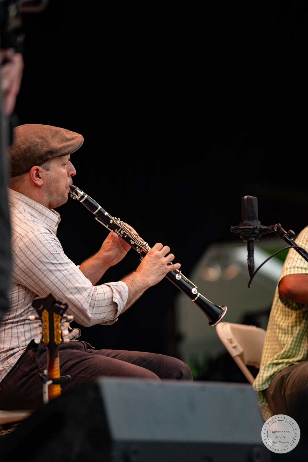 A musician plays clarinet on stage, wearing a flat cap and plaid shirt, performing at an outdoor jazz event.