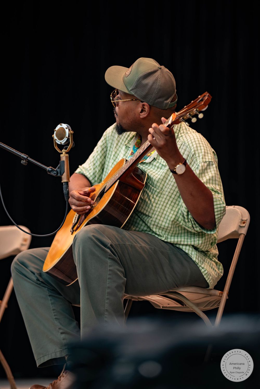 A man in a green plaid shirt and cap plays acoustic guitar while seated on a folding chair near a microphone on stage.