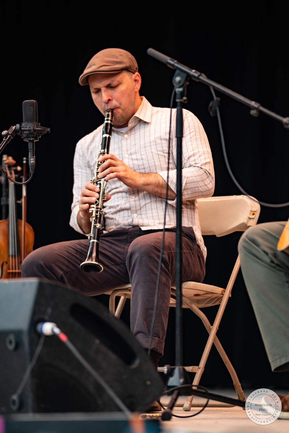 A musician plays clarinet on stage, seated in a folding chair, wearing a brown cap and plaid shirt.