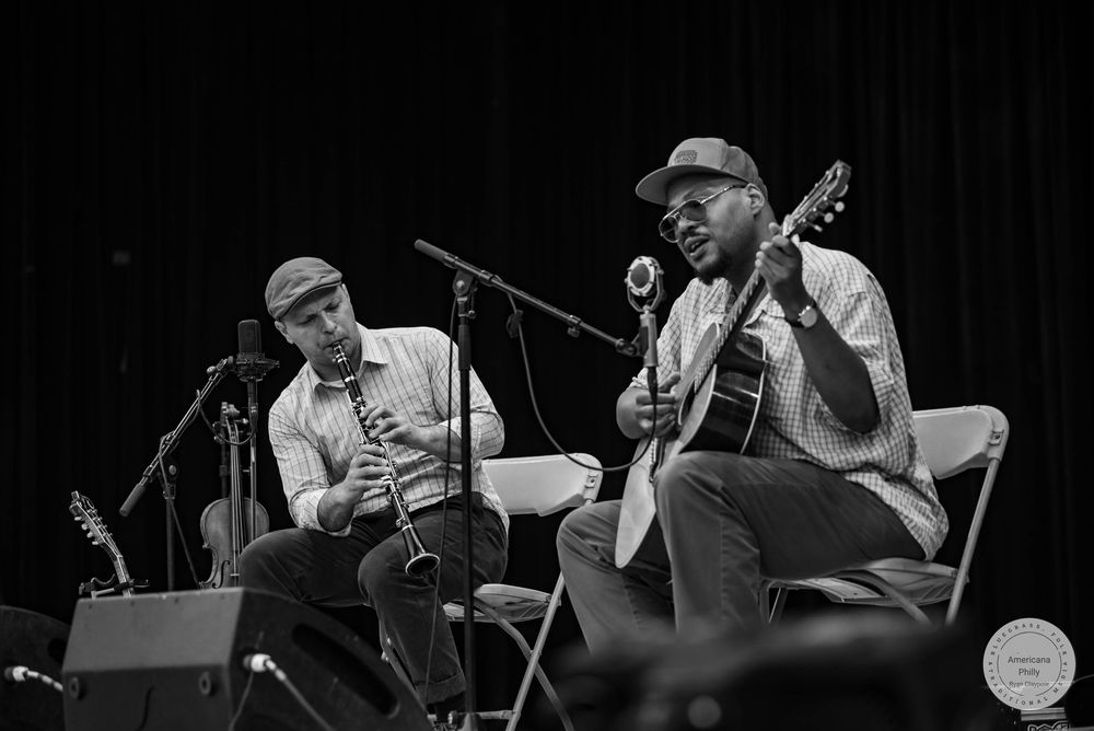 Two musicians perform acoustic guitar and clarinet on stage in black and white photo at live music event.