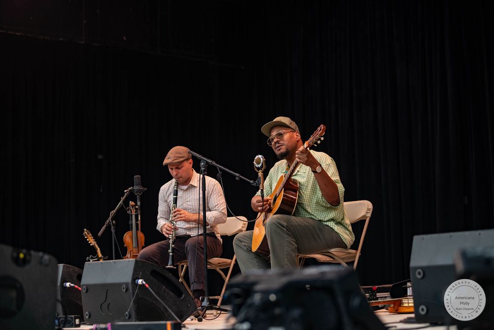 Two musicians perform on stage, one playing clarinet and one playing guitar, seated with microphones at a live music event.