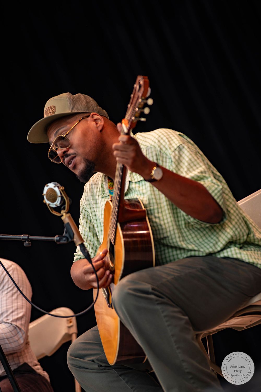 Musician playing acoustic guitar while seated on stage, wearing a green plaid shirt and tan cap.