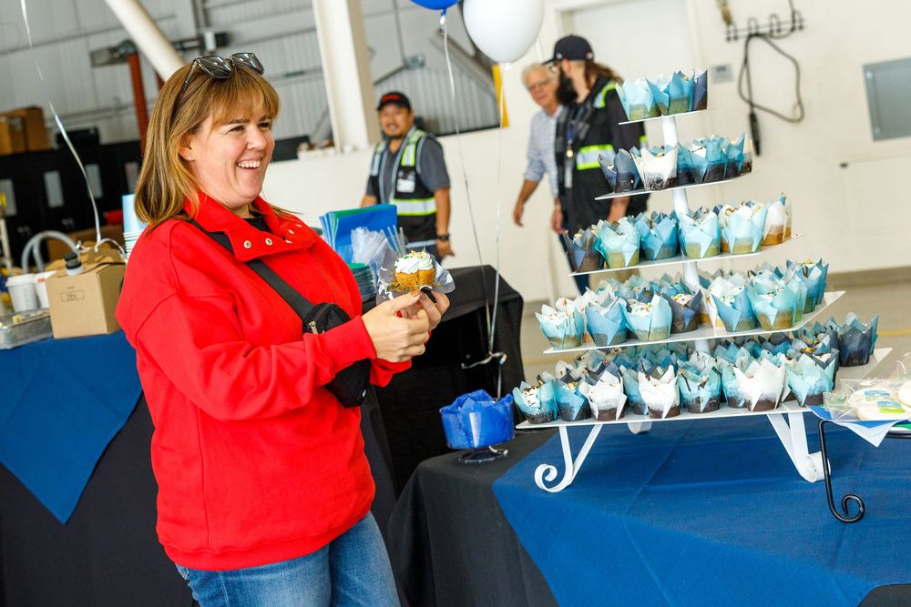 Woman in red jacket holds a drink at an event display table with blue decorative gift bags arranged on a stand.