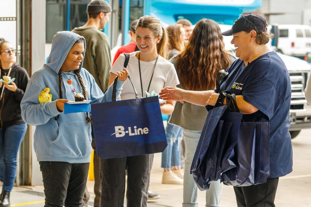 Students receive B-Line branded tote bags outdoors during a school event on a sunny day.