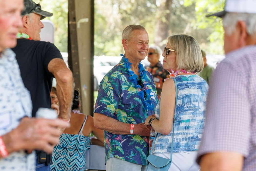 Two adults in tropical shirts socializing outdoors at a casual summer gathering under shaded pavilion.