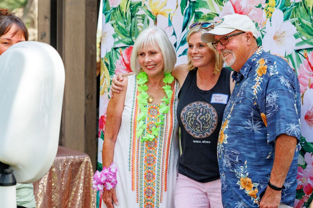 Three friends smile together at a tropical-themed outdoor party, dressed in colorful summer attire.