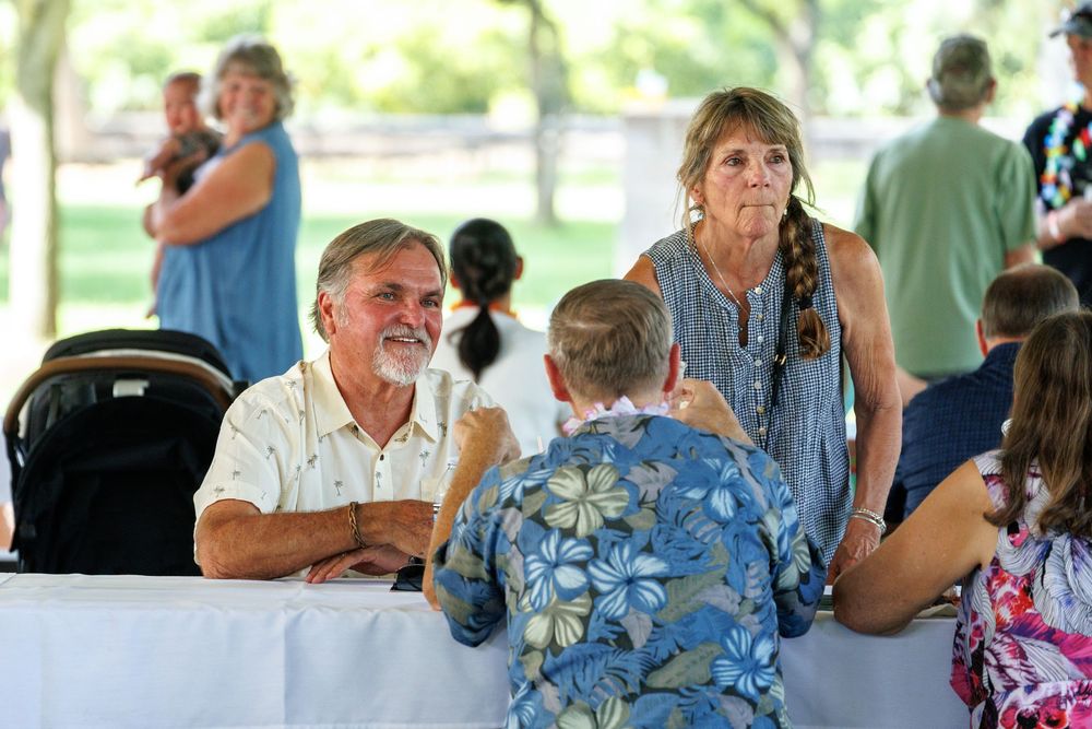 Older man and woman chatting with a child at an outdoor event under a white tent with green trees in background.
