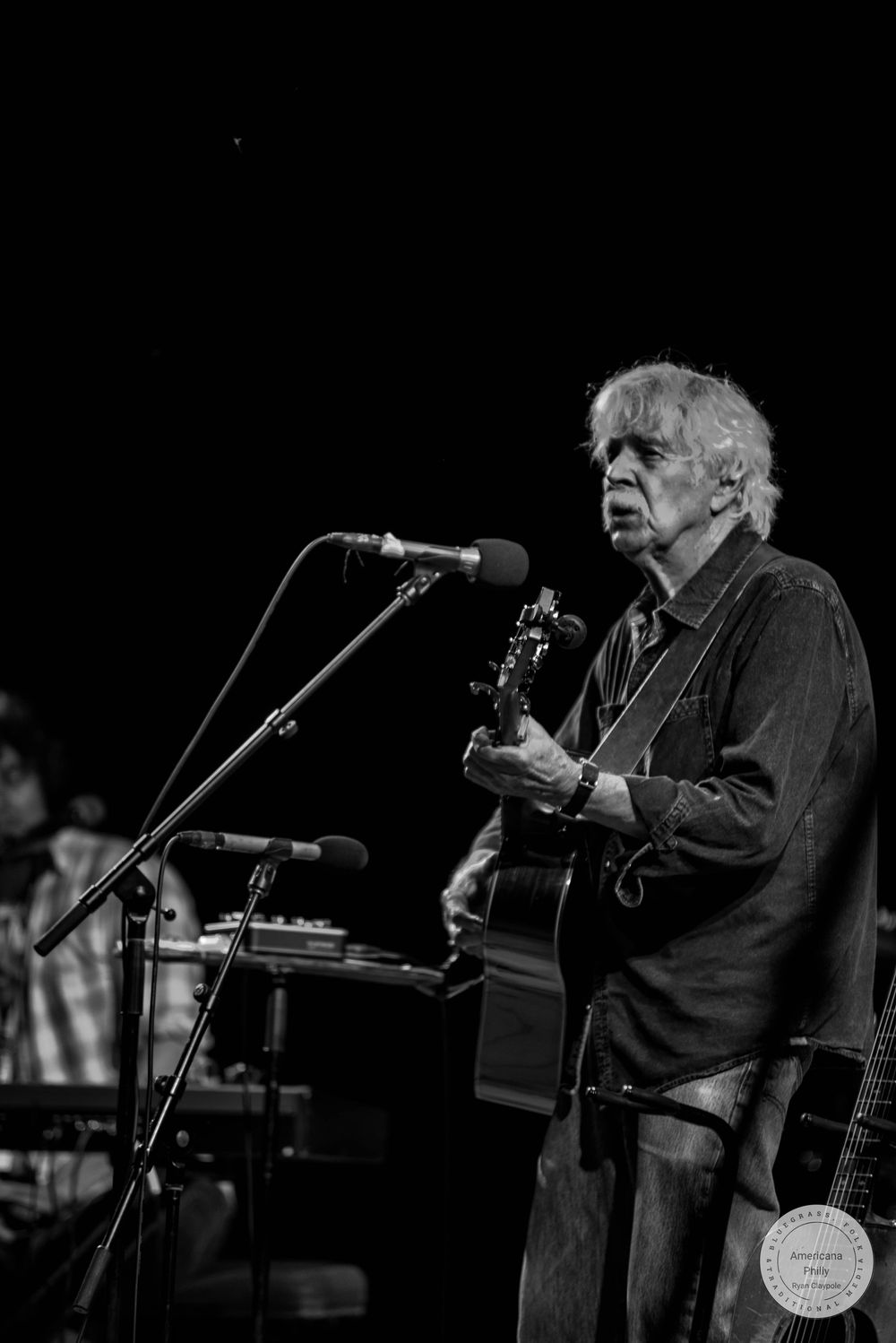 Black and white photo of an older male guitarist performing on stage at a live music concert.