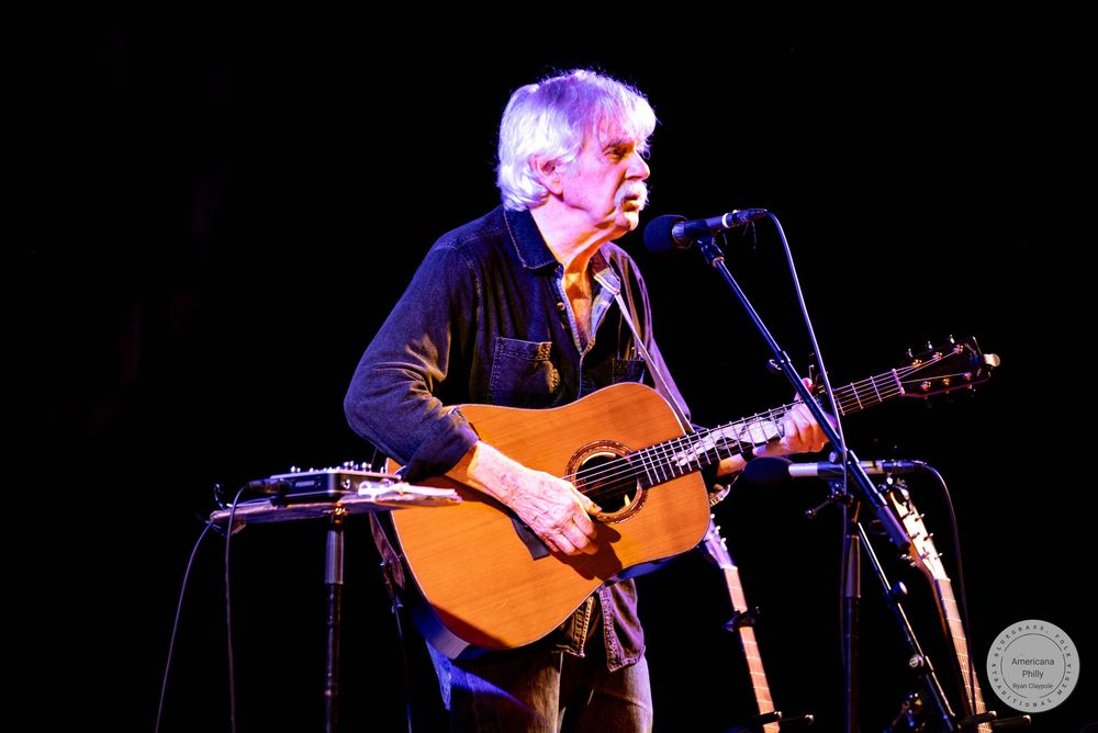 Elderly musician with white hair playing acoustic guitar on stage near microphone in dark concert venue.