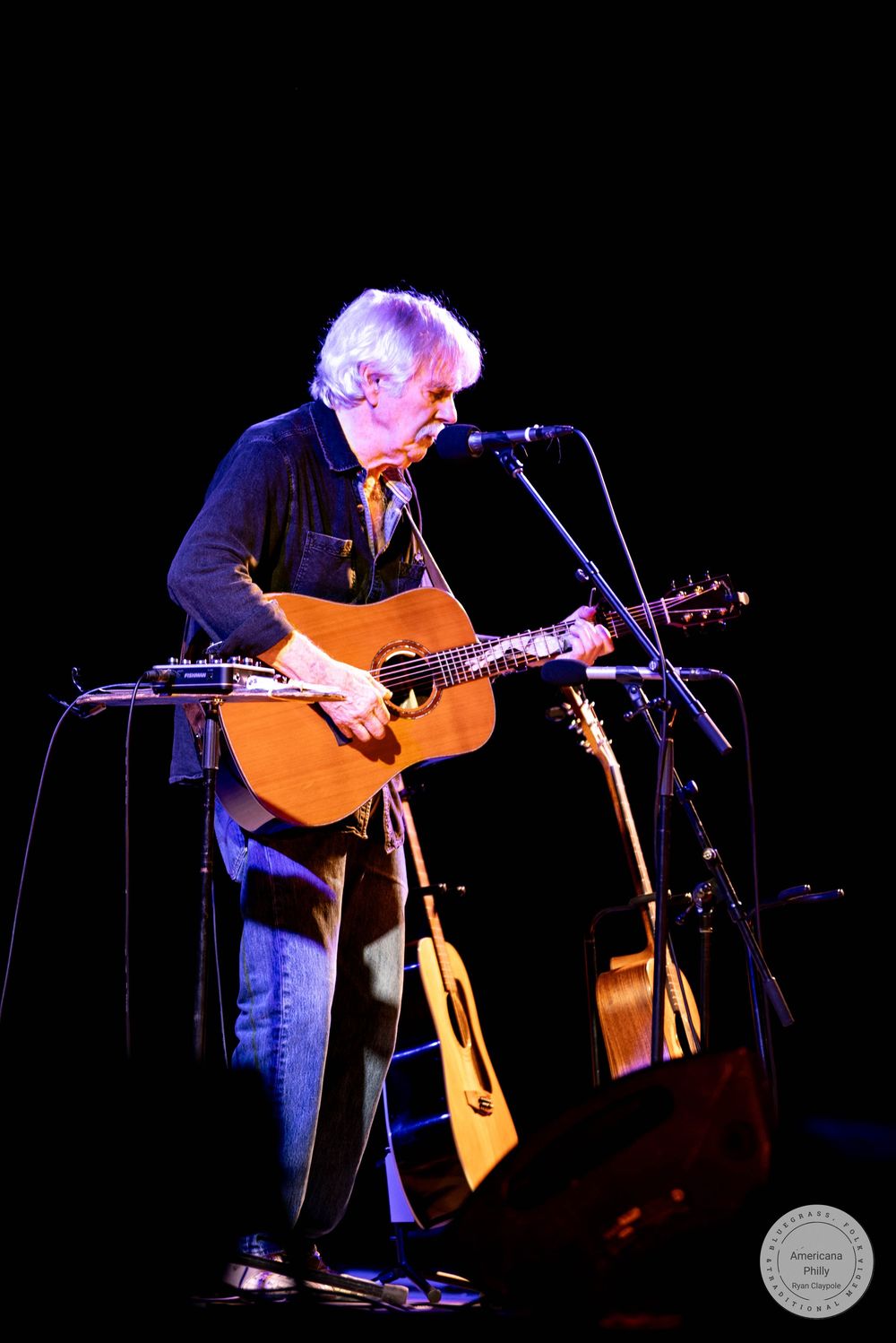 Elderly musician with white hair plays acoustic guitar at microphone stand on dark stage during live performance.