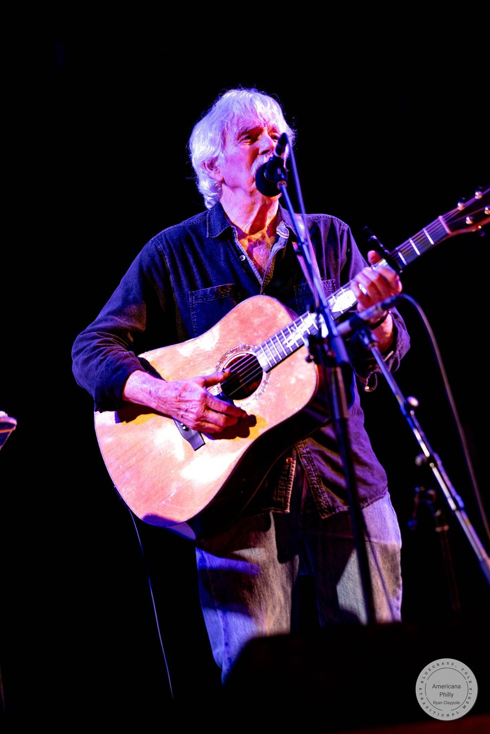 Elderly musician playing acoustic guitar and singing into microphone on dark stage during live performance.