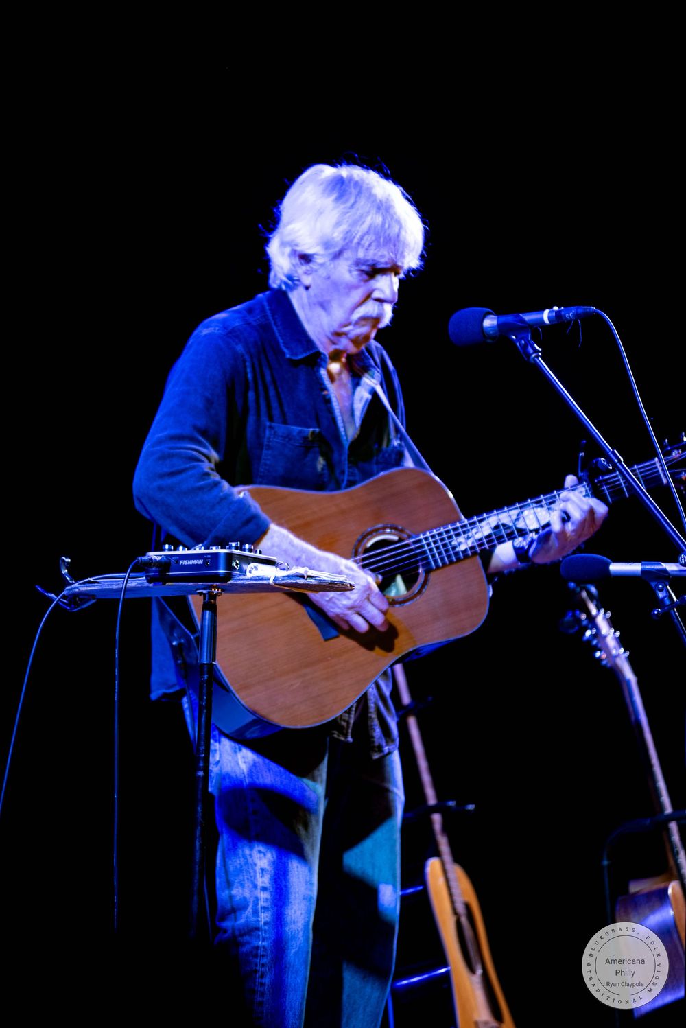 Elderly musician with white hair playing acoustic guitar on stage under blue stage lighting at a live concert.