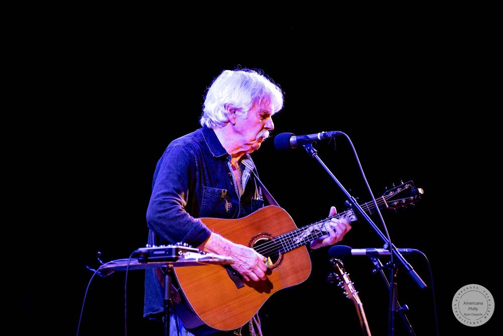Elderly musician with white hair plays acoustic guitar and sings into microphone on dimly lit stage.