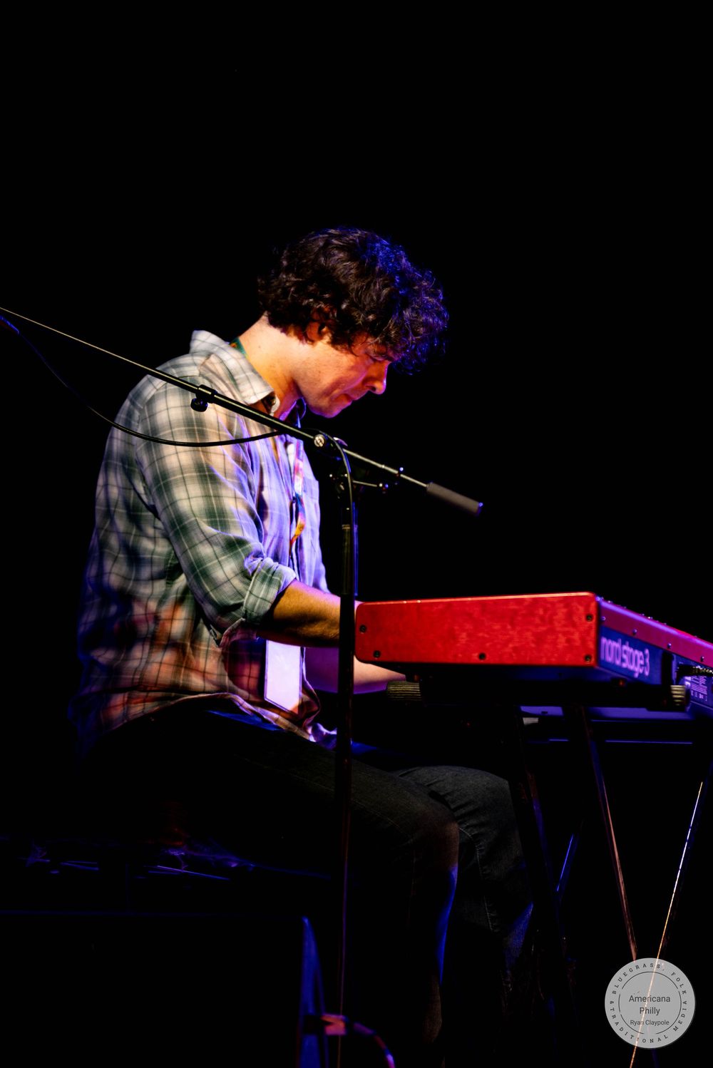 Young man playing a red Nord keyboard on stage, illuminated by warm stage lighting during a live performance.