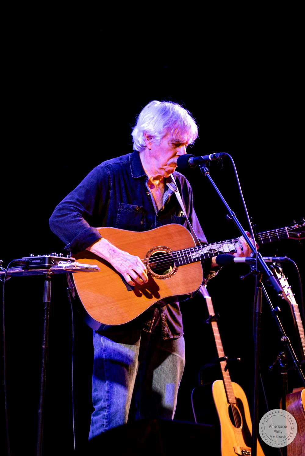 Older musician with white hair playing acoustic guitar on stage at a live concert performance.