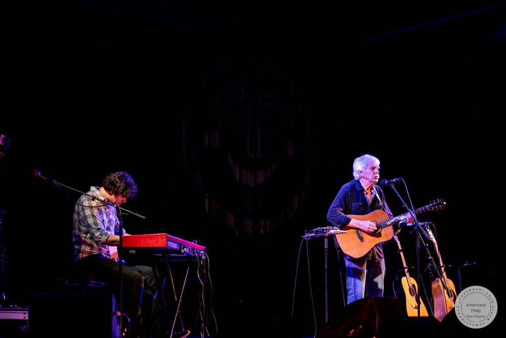 Two musicians perform on stage, one playing keyboard and another on acoustic guitar with microphone in dimly lit venue.