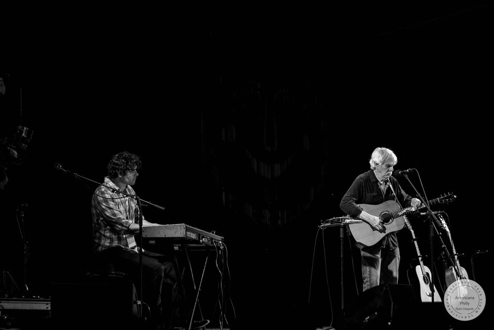 Black and white photo of two musicians performing live, one playing keyboard and one playing acoustic guitar on stage.