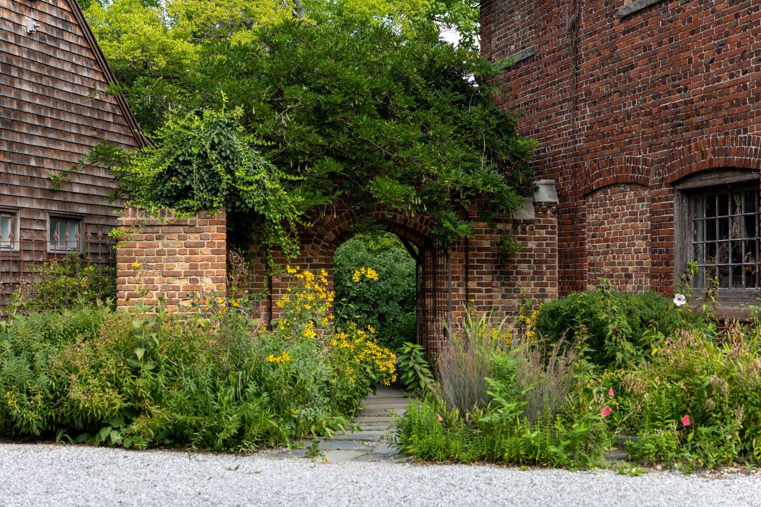 A couple walks hand-in-hand through a lush garden path, dressed in winter coats on a cloudy day.