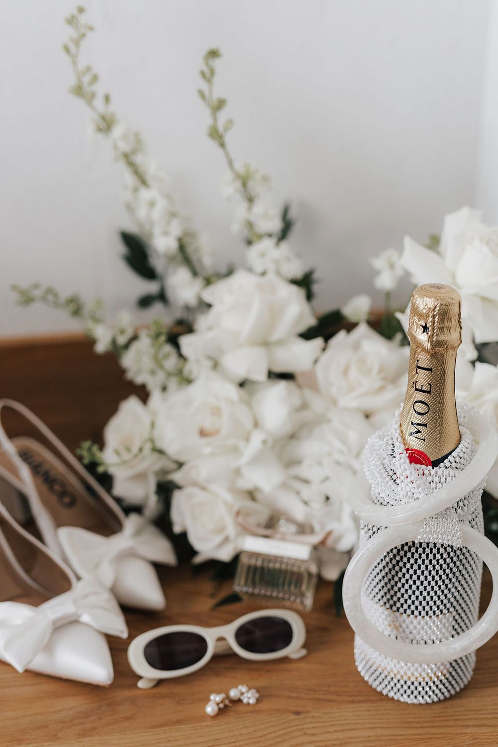 Bridal flat lay with Moet champagne, white roses bouquet, heels, sunglasses, and pearl earrings on wooden surface.