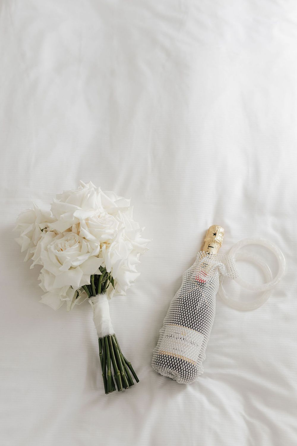 White rose bridal bouquet beside a champagne bottle and rings on white fabric.
