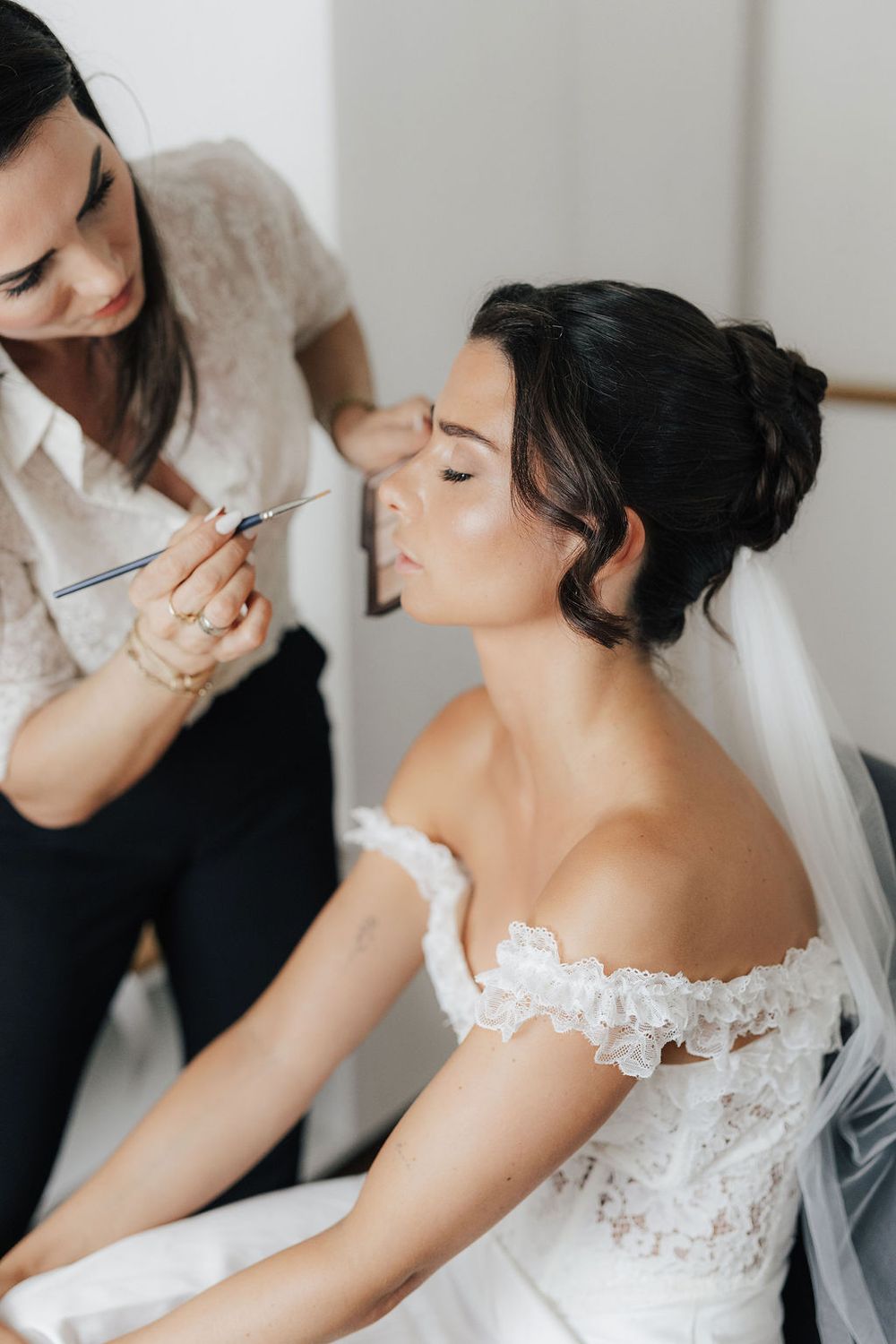 Makeup artist applying eye makeup to a bride in a lace off-shoulder wedding dress before her wedding ceremony.