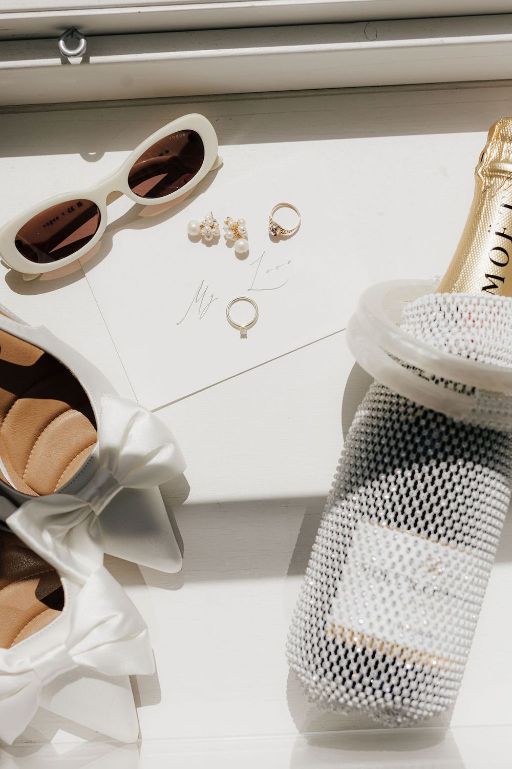 Elegant bridal flat lay with white heels, sunglasses, gold rings, pearl earrings, and Moet champagne in beaded cooler.