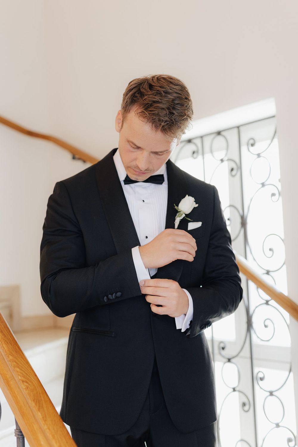 Groom in black tuxedo with bow tie and white rose boutonniere adjusting cufflinks on staircase.