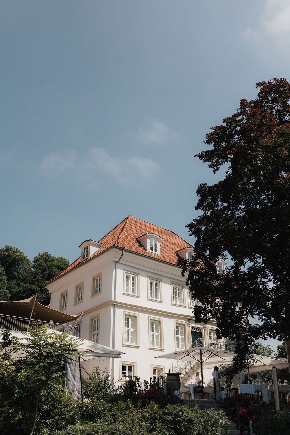 White European villa with orange terracotta roof surrounded by lush trees under a clear blue sky.