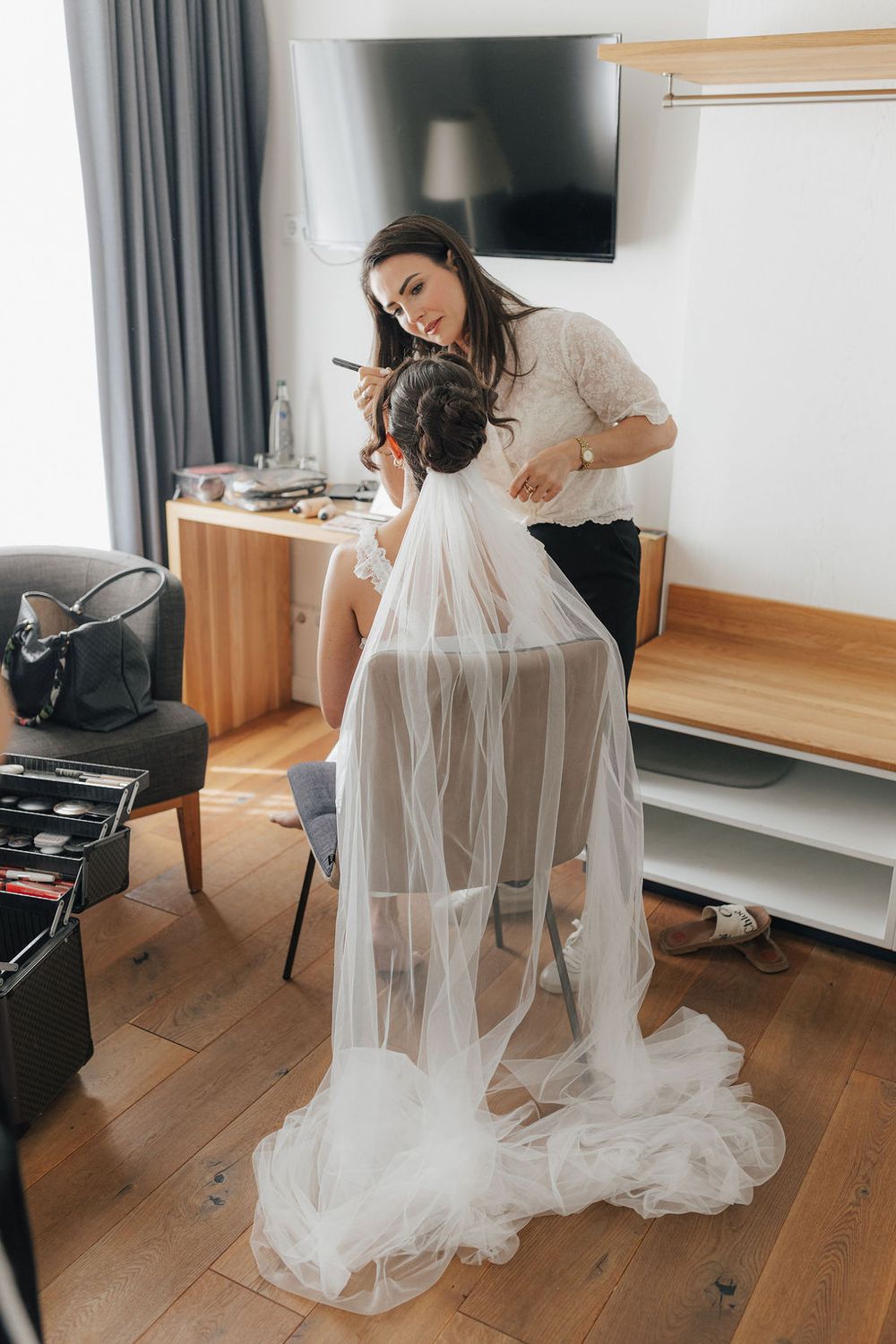 Makeup artist applying makeup to a bride wearing a white veil in a hotel room before the wedding ceremony.