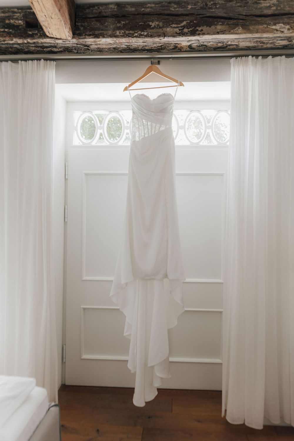 Elegant white strapless wedding dress hanging on a wooden hanger against a bright white door with sheer curtains.