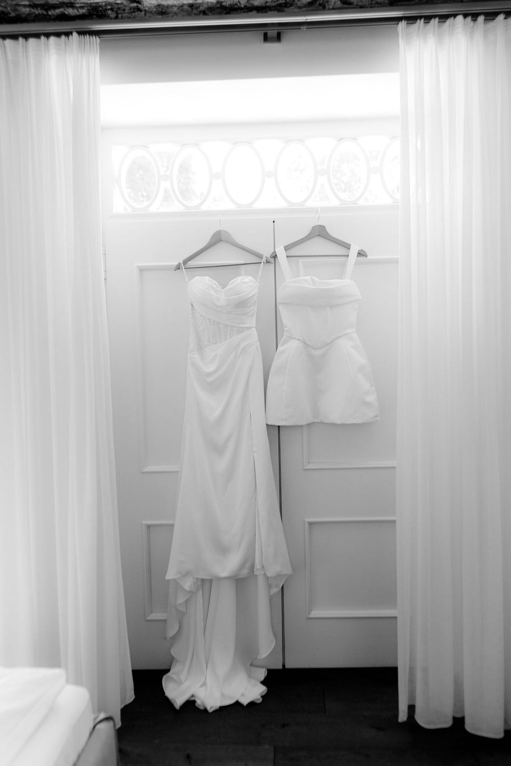 Two white wedding dresses hanging on wooden hangers in a doorway, black and white photography.