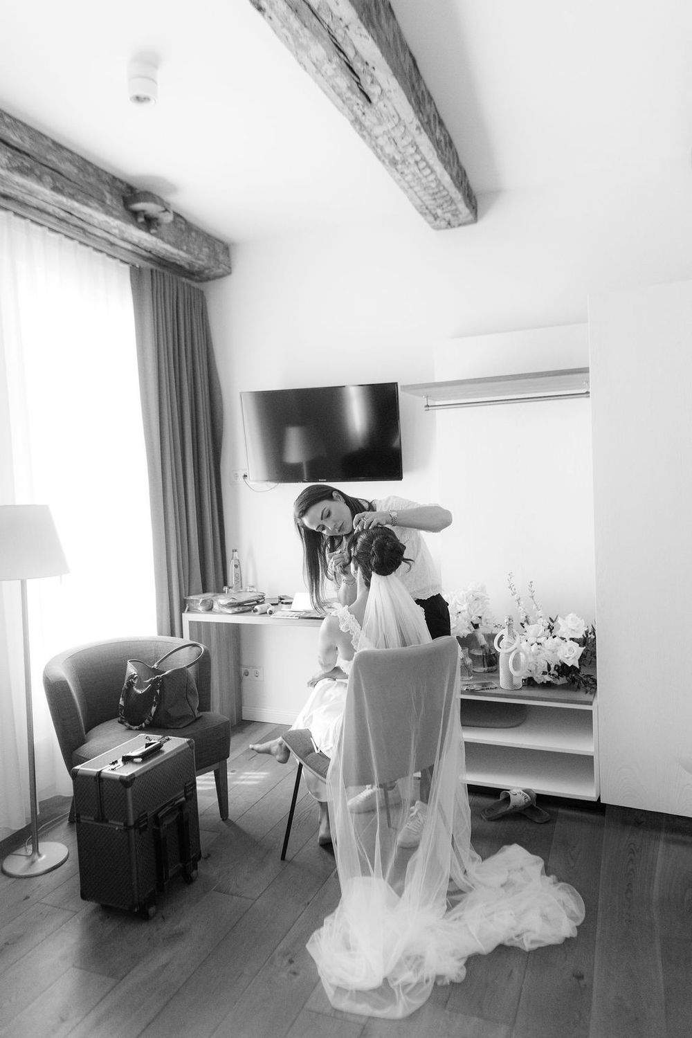 Black and white photo of a bride getting her hair done in an elegant room with wooden beams.