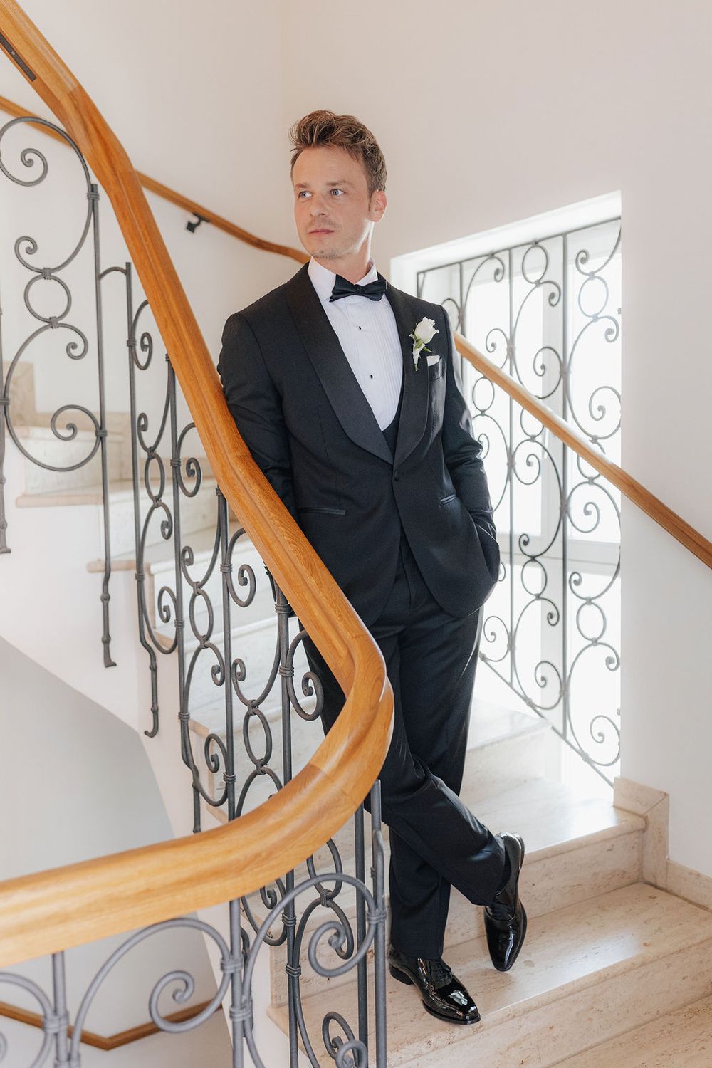A groom in a classic black tuxedo with bow tie poses on an ornate staircase.
