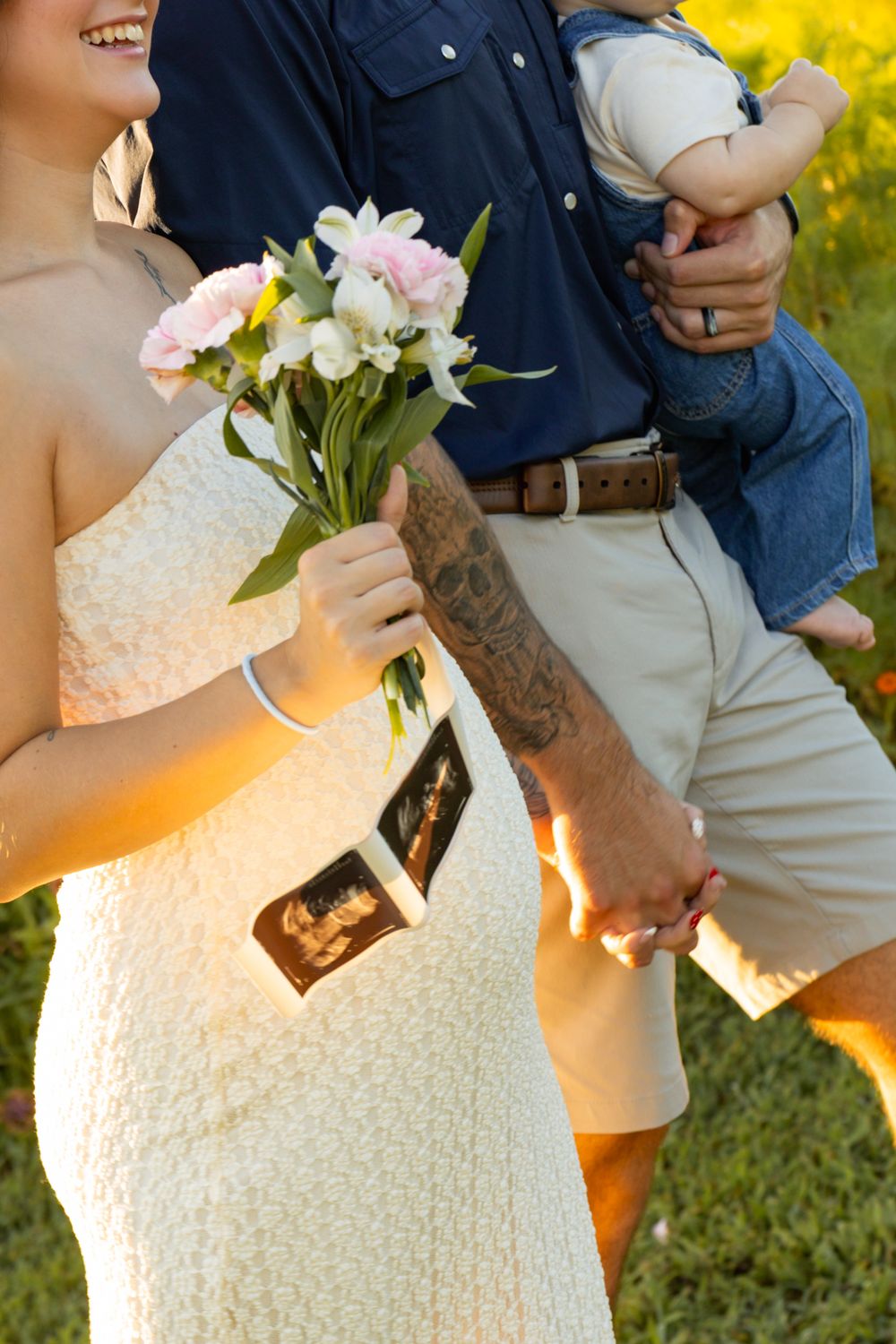 Pregnant woman in white dress holds flowers and ultrasound photos beside partner carrying toddler outdoors.