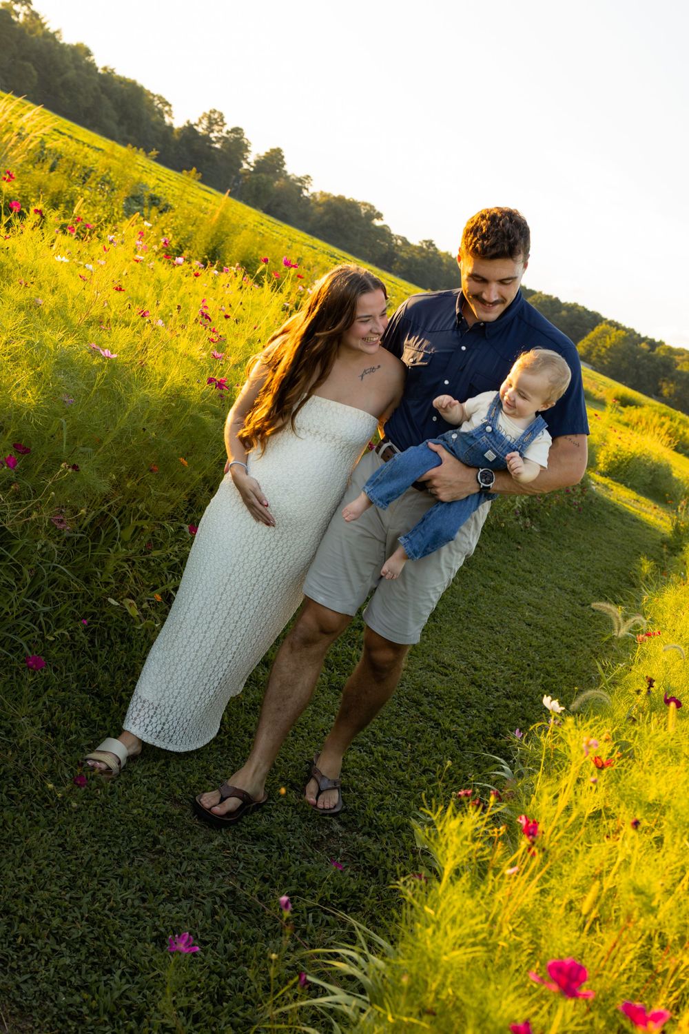 Young family with baby boy poses in a sunlit wildflower meadow, mother in white dress, father in blue shirt.