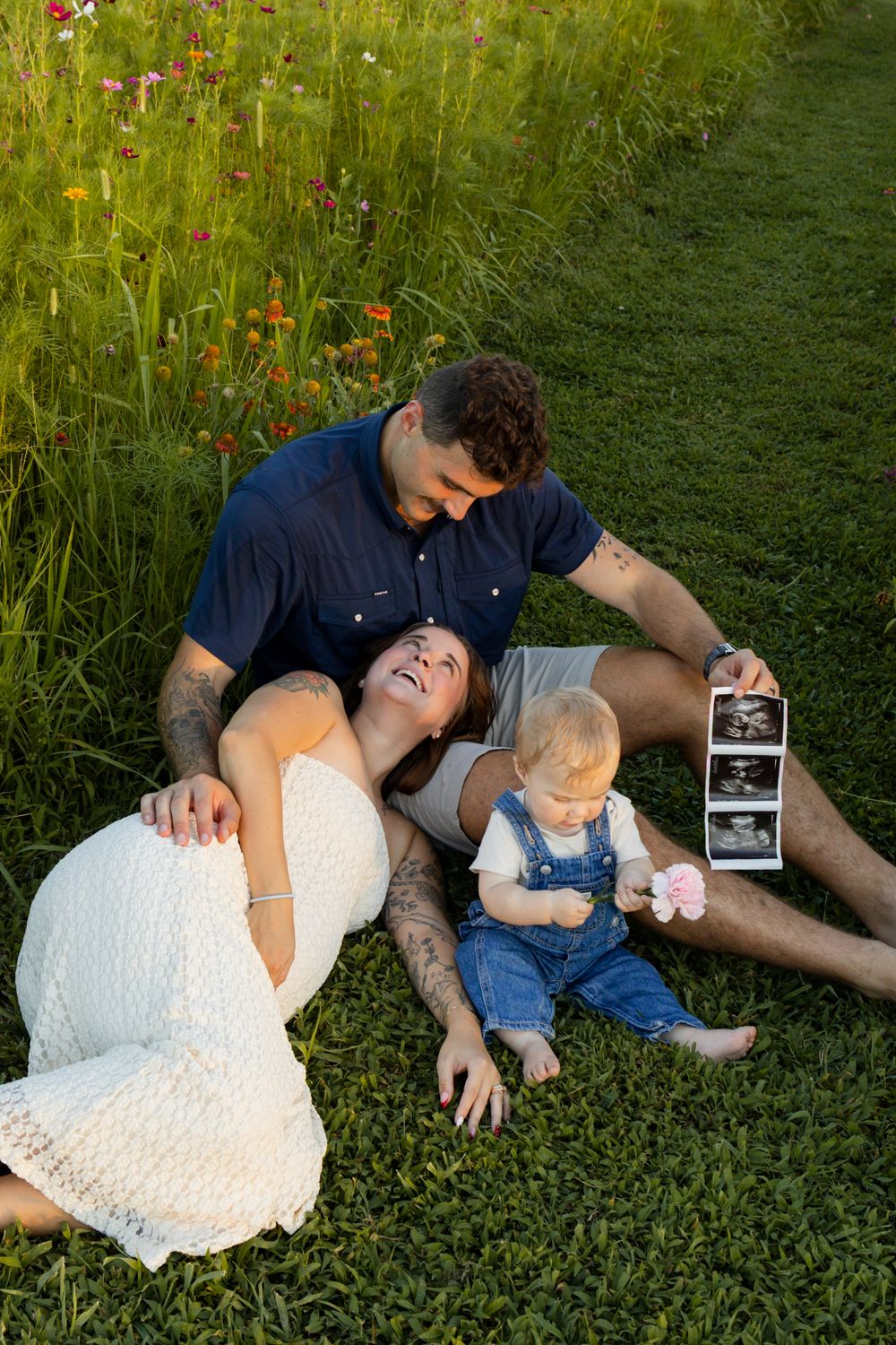 A happy couple sits on grass with their toddler, holding ultrasound photos, announcing a second pregnancy outdoors.