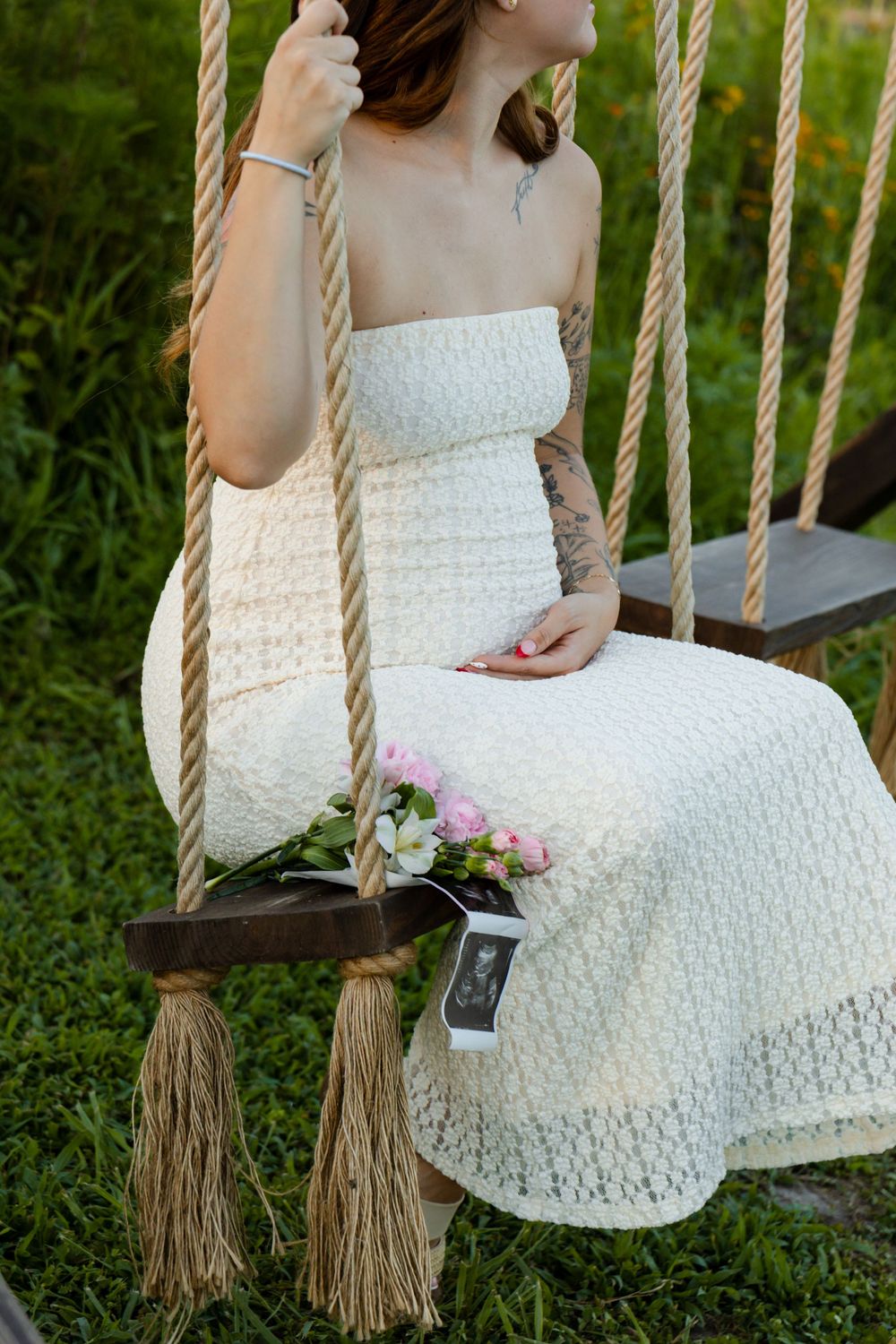 Pregnant woman in white lace dress sitting on rope swing outdoors, holding flowers and ultrasound photo.