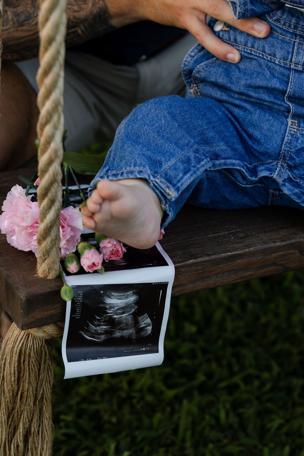 Pregnant woman on rope swing with ultrasound photo and pink flowers, wearing denim overalls outdoors.