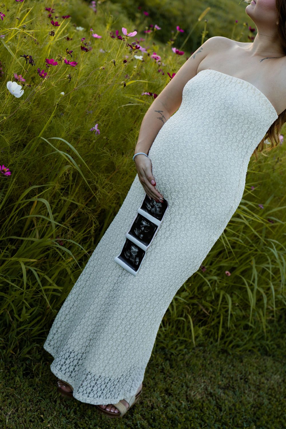 Pregnant woman in white strapless dress holding ultrasound photos in a lush green flower field.