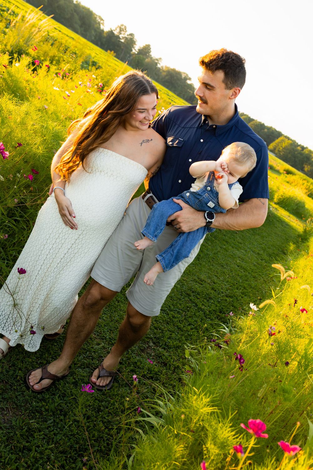 Young family with baby boy poses in a sunny wildflower field, mom in white dress, dad in blue shirt.