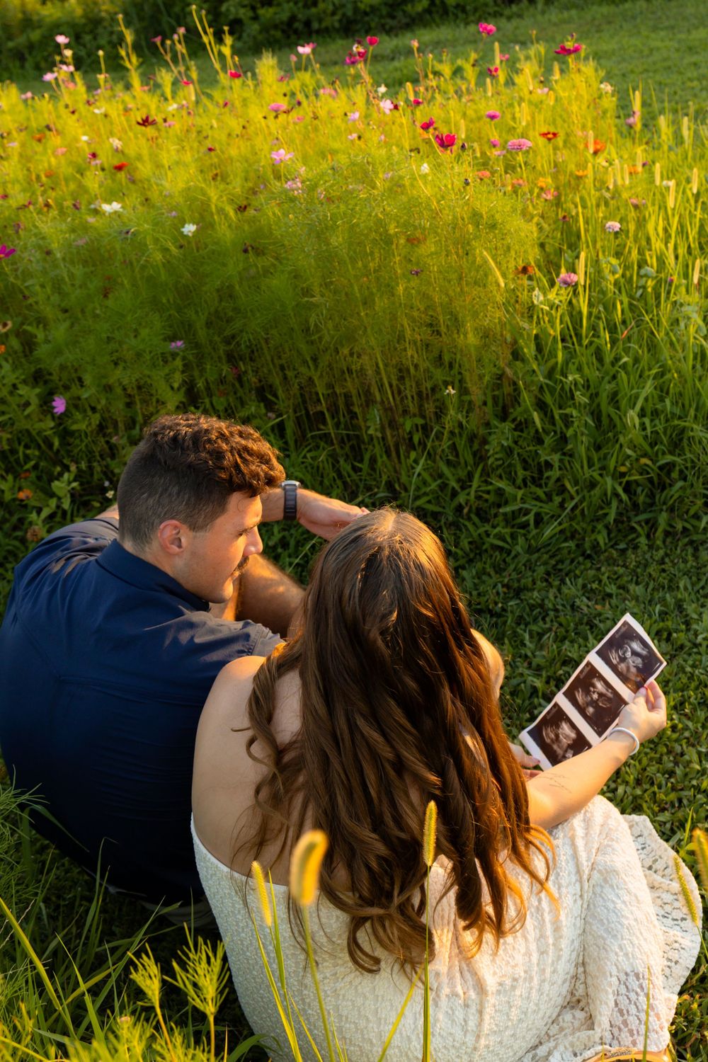 Couple sitting in a sunny garden looking at ultrasound photos surrounded by colorful wildflowers.