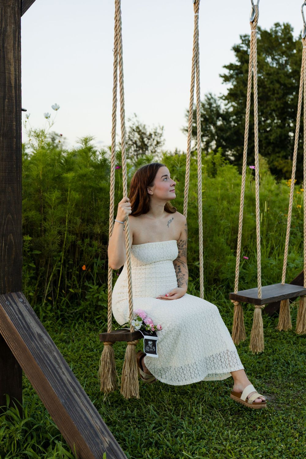 Pregnant woman in white dress sitting on rope swing outdoors, holding ultrasound photo, surrounded by lush greenery.