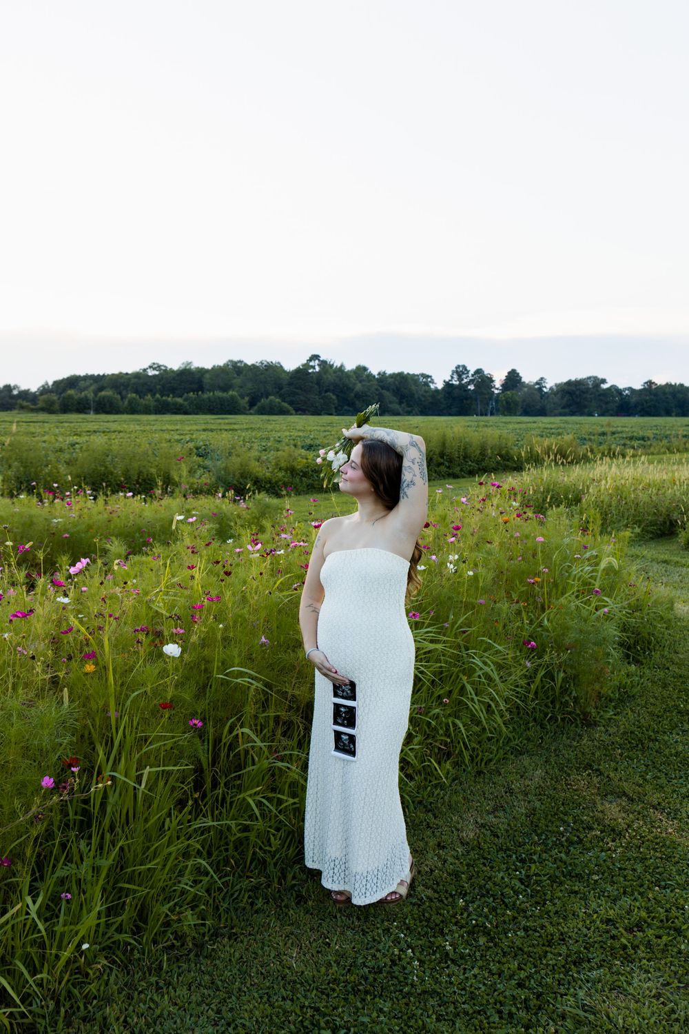 Pregnant woman in white strapless dress holding ultrasound photos in a lush green flower field at dusk.