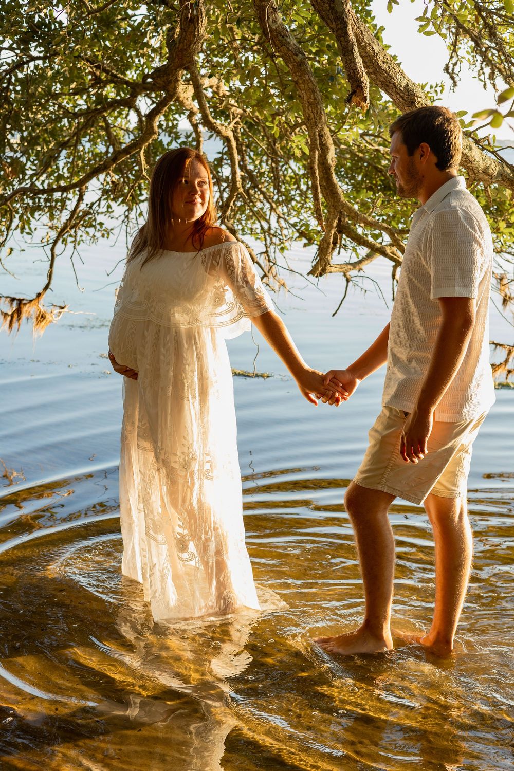 Pregnant woman in white dress holds hands with man standing in shallow water under tree at golden hour.