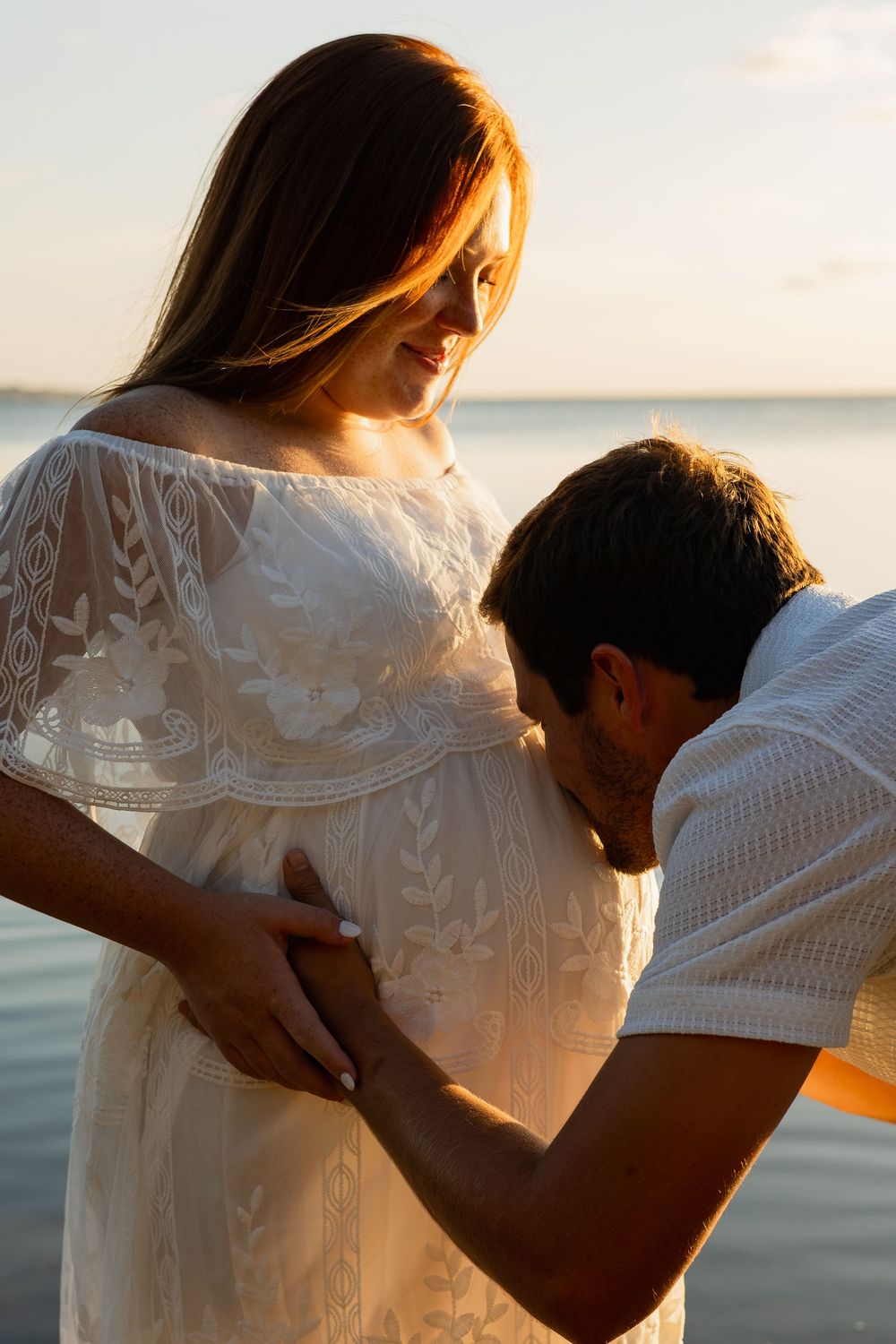 A man kisses a pregnant woman's belly at sunset by the water, as she smiles in a white lace dress.