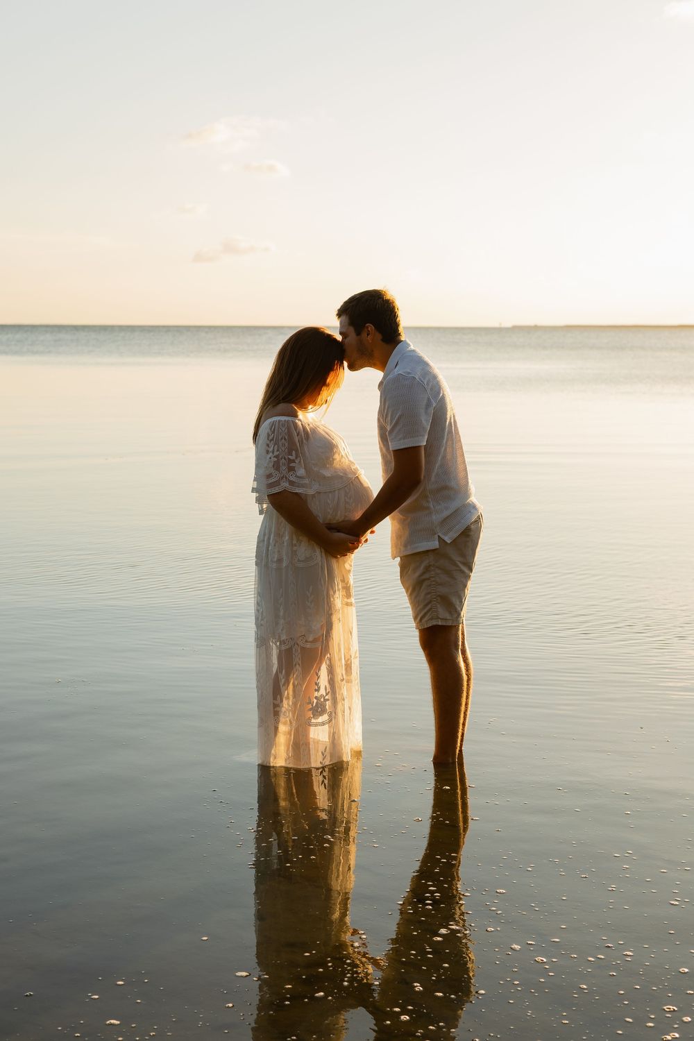 Pregnant couple standing in shallow ocean water at sunset, sharing an intimate moment in white outfits.